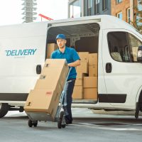 Delivery Man Pushes Hand Truck Trolley Full of Cardboard Boxes H Delivery Man Pushes Hand Truck Trolley Full of Cardboard Boxes Hands Package to a Customer.
