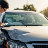 A woman closely examines and writes notes on the damage to the front of a silver car after an accident during daylight on an urban street.