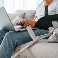 Sitting on the sofa with laptop. Man with crutches is at home indoors. Having injuries.