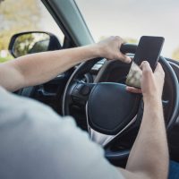 Young driver guy distracted by his phone while in front of the steering wheel, using his smartphone with one hand while driving. Risk and danger situations on the road, violating traffic rules