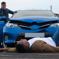 A man is lying in front of a damaged car which has just hit him. The responsible driver is standing at the door of the car, looking at him with a worried expression.