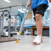 Medical care patient walking using crutches to support her injured ankle sprain, in the hospital physical therapist room.