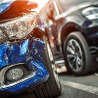 Damaged blue car front after collision parked next to a black vehicle during daylight in urban parking lot with visible dents and scratches on bumper and headlight